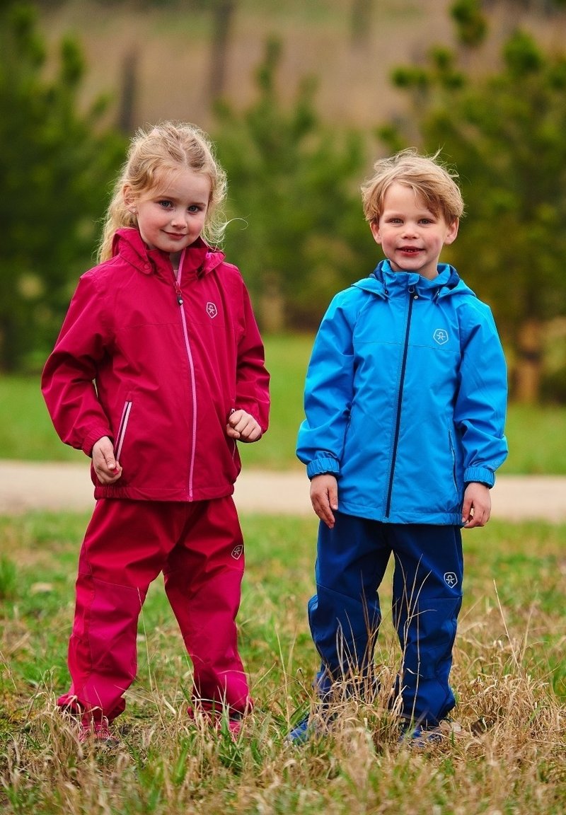 Costume de ploaie pentru copii, în roșu și albastru, realizate din material impermeabil. Fiecare are glugă, fermoar în față și manșete elastice, stând pe iarbă.