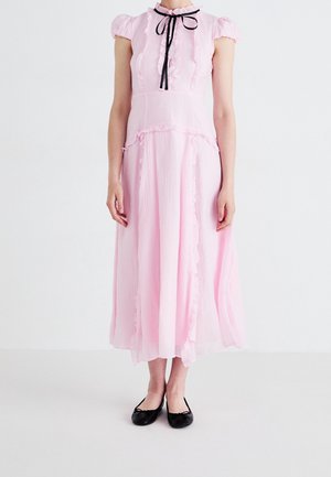 Nainen pukeutuneena vaaleanpunaiseen, röyhelöiseen midimekkoon, jossa on lyhyet hihat ja musta nauhavyö, yhdistettynä mustiin balleriinakenkiin.