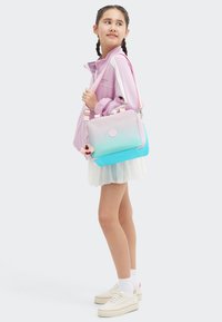 Borsa con sfumature rosa e blu, dalla texture liscia, dotata di accessori in argento e un soffice charm rosa, portata da una ragazza con giacca rosa e gonna bianca.
