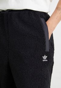 Zwarte fleecesweatpants hebben een zachte, gestructureerde oppervlakte, zijzakken met zwarte stofafwerking en een wit adidas-logo aan de voorkant.