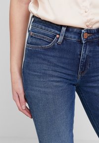 Jeans i denim med en medelblå till mörkblå gradient, med hög midja, fem fickor och detaljer i kopparfärg.