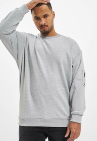 Grauer Pullover mit langen Ärmeln, geripptem Rundhalsausschnitt und einer Reißverschlusstasche am linken Ärmel. Weiches, strukturiertes Material.