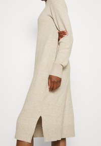 Beige stickad klänning med långa ärmar, ribbade muddar och sidofickor, tillverkad av ett mjukt, strukturerat tyg.