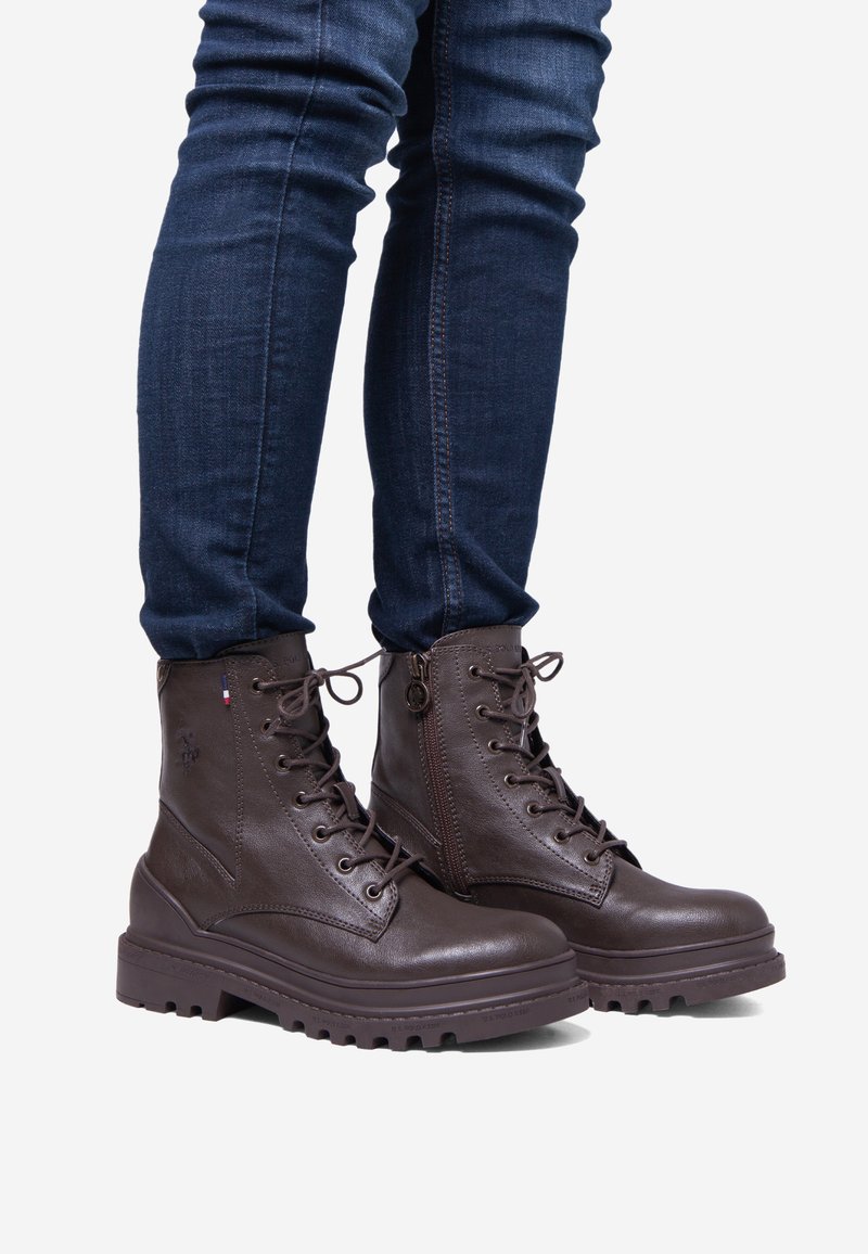 Botas de combate de cuero marrón con una suela robusta, frente con cordones, cremallera lateral y detalles de costura. Combinadas con jeans ajustados de color azul oscuro.