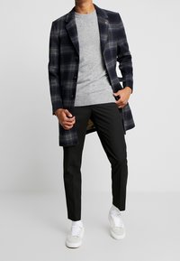 Abrigo de cuadros oscuros con cuello de muesca, suéter de punto gris, pantalones negros y zapatillas blancas. El abrigo cuenta con un cierre de botones y bolsillos.