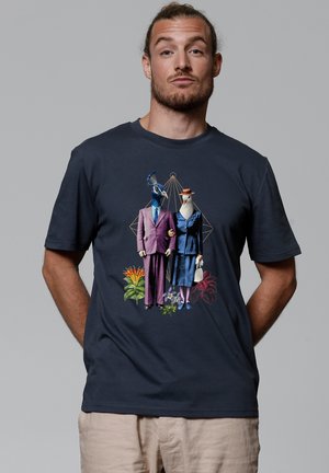 Zwarte t-shirt met een graphic van twee vogels in formele kleding, omringd door kleurrijke bloemen. De mannelijke vogel in een paarse pak, de vrouwelijke in blauw.