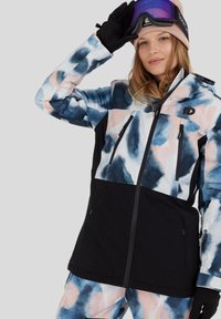 Skijas met een meerkleurig aquarelpatroon in blauw en roze, zwarte panelen, ritssluitingen en verstelbare capuchon. Draagt handschoenen en een skibril.