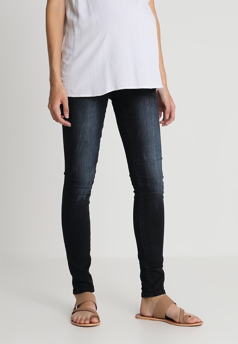 Personne portant un jean skinny foncé, un haut blanc et des sandales plates beiges, debout devant un fond clair uni.