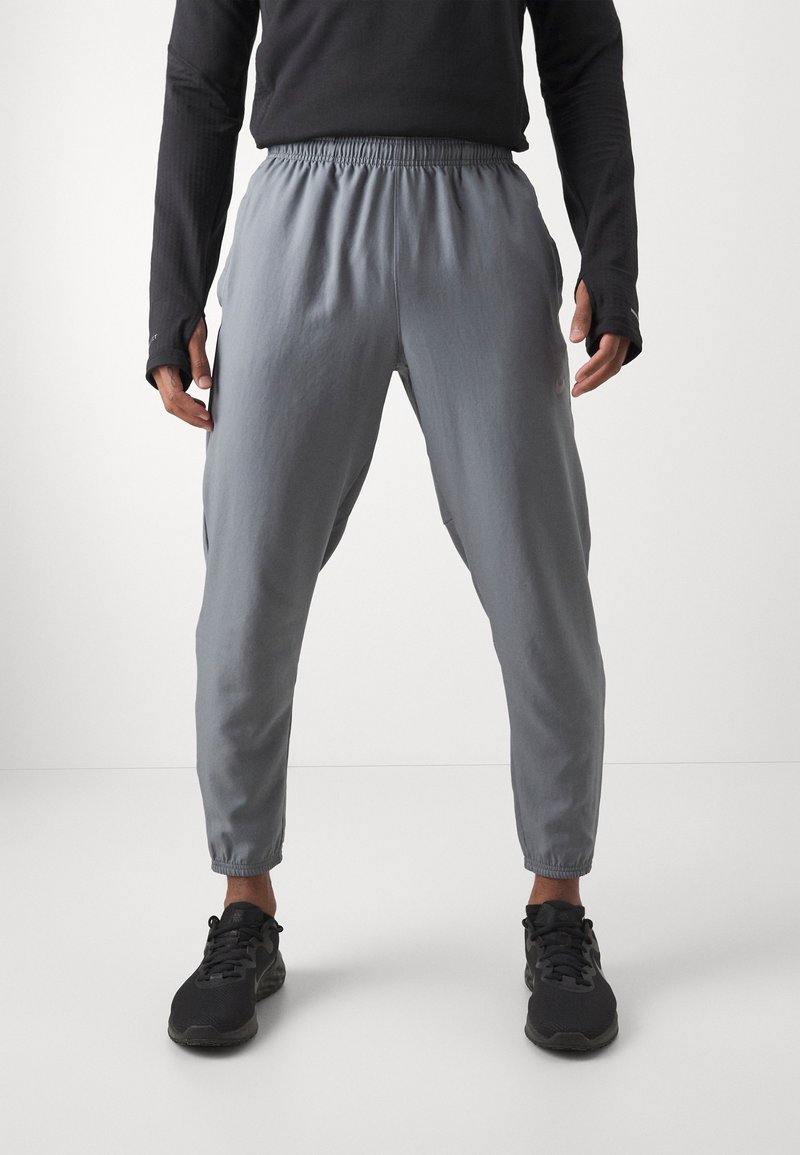 Personne portant un pantalon de jogging gris tapered avec une ceinture élastique et des chaussures de sport noires, debout devant un fond clair uni.