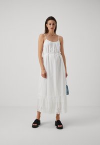 Balta maksi kleita ar plānām siksnām, saistītu vidukli, volānu apakšmalu un acs apdari. Modelis valkā melnas platformas sandales.
