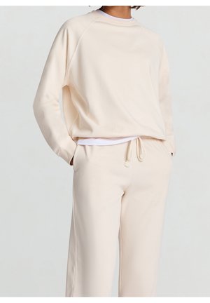 Person iført cremefarvet sweatshirt og matchende joggingbukser med snøre i lommerne mod en ensfarvet baggrund.