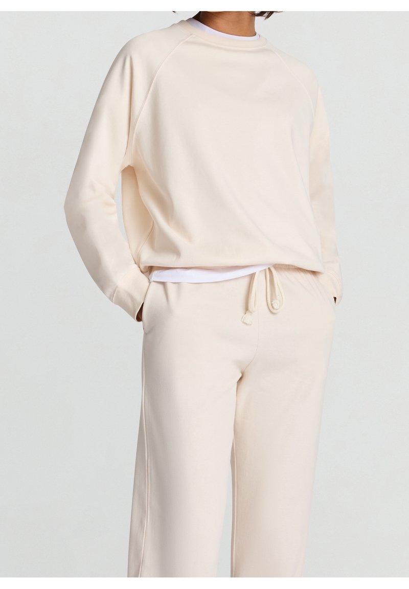Personne portant un sweat-shirt crème et un pantalon de jogging assorti avec cordon de serrage, les mains dans les poches, sur un fond uni.