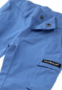 Vaaleansiniset lasten housut, jotka on valmistettu puuvillasekoitteesta. Housuissa on napitus, tasku jalassa ja musta "Villervalla" -merkki.