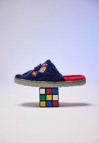 Zapatilla deslizante de color azul marino y peluda con acentos rojos, parches bordados y una suela de goma texturizada, exhibida sobre un cubo Rubik.
