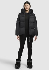 Black puffer jacket with a high collar, featuring puffed sections and a front zip. Worn with black leggings and glossy black boots.