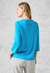 Türkises Langarmshirt mit rundem Halsausschnitt und drei Viertelärmeln. Mit einem kleinen Logodetail auf der Rückseite. Glatte Stofftextur.
