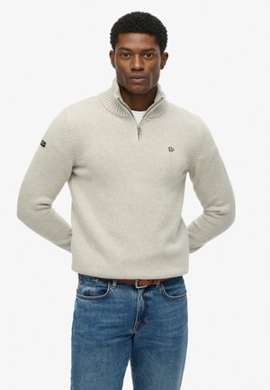 Suéter gris claro con cuello con cremallera, textura acanalada y emblema en el pecho. Combinado con jeans azules y un cinturón marrón.