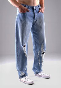 Helle blaue, zerfledderte Denim-Jeans mit großen Löchern, ausgefransten Kanten und lockerer Passform, kombiniert mit weißen Segeltuch-Sneakern.