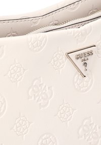 Superficie de cuero blanco texturizado con patrones florales en relieve y una placa metálica triangular del logo de Guess USA cerca de una cremallera.