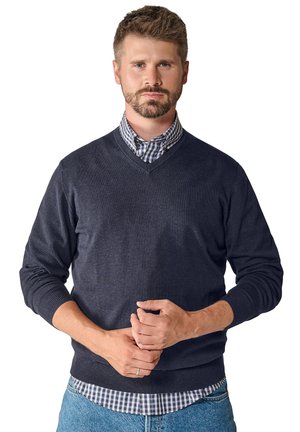 Pullover aus dunkelblauem Wollmix mit V-Ausschnitt. Das Model trägt ein blaues und weißes kariertes Hemd darunter, kombiniert mit hellblauen Jeans.