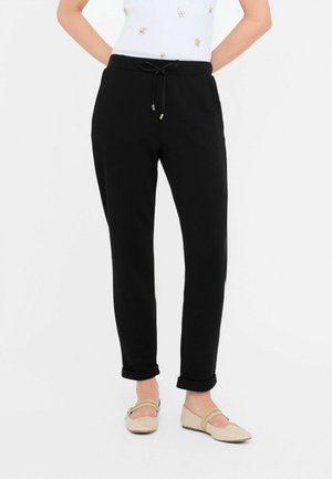 Zwarte, taps toelopende joggingbroek met een drawstring waist, gladde textuur, cuffed enkels en zijzakken, gedragen met lichtbeige platte schoenen.
