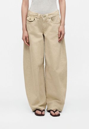 Person iført løse, beige bukser med brede ben, en lysegrå top og mørke sandaler, stående mod en ensfarvet hvid baggrund.