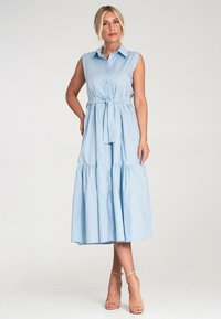 Jasnoniebieska sukienka bez rękawów z guzikiem z przodu, zaciśnięty pas z paskiem i falbanistym dołem. Gładki bawełniany materiał, długość do kolan.