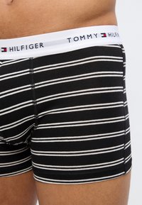 Ανδρικός κορμός που φοράει μαύρα και άσπρα ριγέ μποξέρ Tommy Hilfiger με λευκή ζώνη μέσης που φέρει το λογότυπο και το κείμενο της μάρκας.