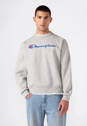 Junger Mann trägt ein graues Champion-Sweatshirt mit blauem Logo und hellblauen Jeans, steht vor einem schlichten weißen Hintergrund.