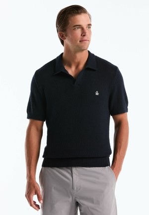 Mann trägt ein kurzärmeliges schwarzes Strick-Poloshirt mit kleinem Pinguin-Logo, kombiniert mit hellgrauen Hosen, steht mit einer Hand in der Tasche.