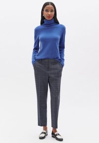Pull à col roulé bleu associé à un pantalon à carreaux gris. Le pantalon est ajusté avec une coupe droite. Des mocassins noir et blanc complètent le look.