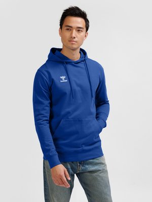Homme portant un sweat à capuche bleu avec le logo « hummel », debout, une main dans la poche et regardant sur le côté.