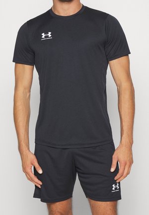 Homme portant un t-shirt de sport noir à manches courtes Under Armour et un short assorti, debout devant un fond clair uni.