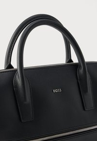 Mala preta em couro texturizado, com duas pegas, um fecho de correr prateado e um pequeno logótipo metálico. Design compacto e estruturado.