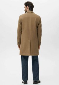Abrigo de lana marrón con un diseño entallado, con una abertura sencilla en la parte posterior, combinado con pantalones de mezclilla oscuros y zapatos negros. Fondo neutro.