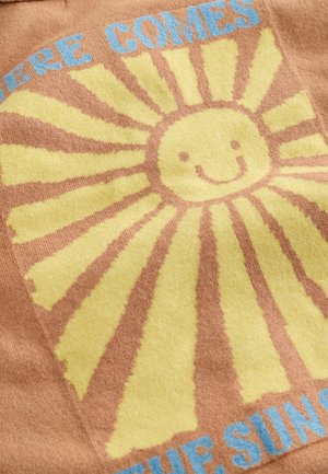 Uśmiechnięte żółte słońce z promieniami na brązowej tkaninie, częściowo z niebieskim napisem "HERE COMES" oraz "THE SUN".