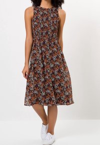 Robe midi sans manches avec un motif floral orange, blanc et vert sur fond sombre, portée avec des baskets blanches, modèle debout avec une jambe croisée.