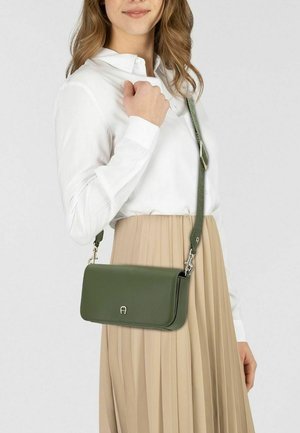 Borsa a tracolla in pelle verde con design a pattina, ferrature argento e tracolla regolabile, indossata sopra una gonna beige plissettata e una camicia bianca.