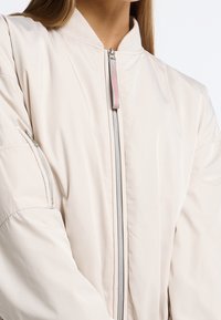 Primo piano di una persona che indossa una giacca bomber beige chiaro con zipper frontale e tasca con zip sulla manica, in piedi contro uno sfondo neutro.