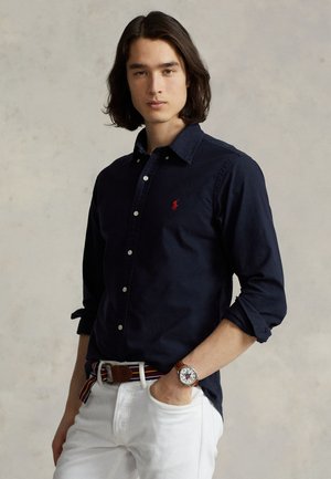 Jeune homme aux cheveux mi-longs portant une chemise bleu marine, un pantalon blanc, une ceinture rayée et une montre-bracelet, debout devant un fond uni.