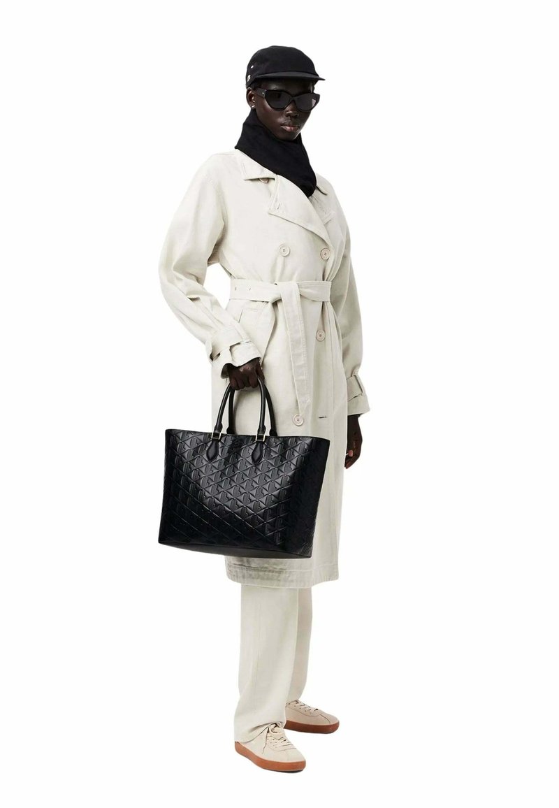 Sac de toile matelassé noir avec doubles poignées, mettant en avant une finition brillante. Associe à un trench-coat beige et des baskets claires.