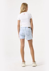 Haut court à rayures blanches avec logo rouge au dos, associé à un short en denim bleu clair. Le mannequin porte des baskets beiges. Look simple et décontracté.
