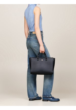Personne en haut bleu sans manches et jean tenant un grand sac à main en cuir noir avec doubles poignées, debout devant un fond uni.