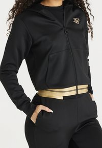 Zwarte ritsjas met een gouden logo, voorzien van een capuchon en zakken aan de voorkant. Gecombineerd met hoge taillebroeken, geaccentueerd door een gouden riem.