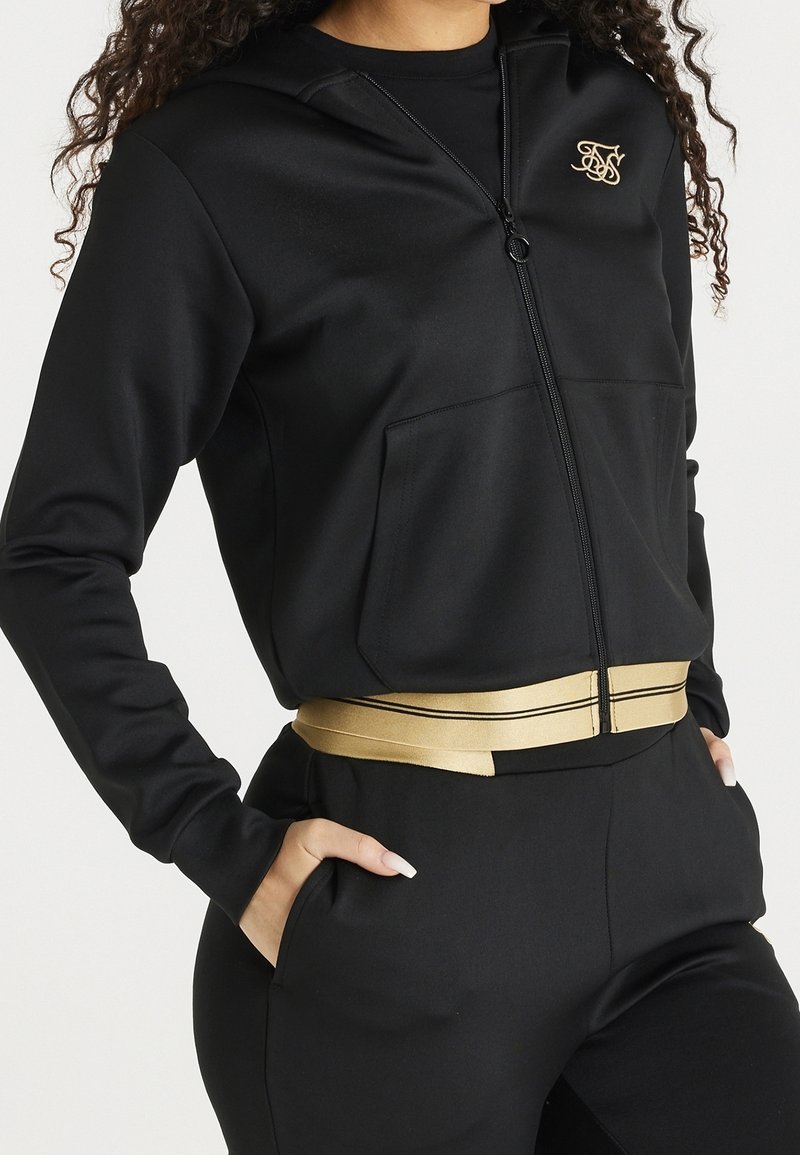 Zwarte ritsjas met een gouden logo, voorzien van een capuchon en zakken aan de voorkant. Gecombineerd met hoge taillebroeken, geaccentueerd door een gouden riem.
