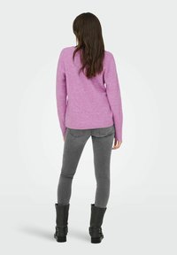 Femme debout, vue de dos, portant un pull rose, un jean skinny gris et des bottes noires mi-mollet, sur un fond clair uni.