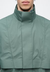 Svetlozelená bunda s vysokým golierom, hladkou textúrou a čistými líniami; má minimalistický dizajn a štruktúrovanú siluetu.