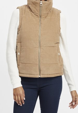 Beżowa ocieplana kamizelka sztruksowa z zamkiem błyskawicznym z przodu i kieszeniami na zatrzaski, noszona na białej prążkowanej bluzce z długim rękawem oraz ciemnoniebieskich dżinsach.