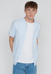 Jonge man met kort krullend haar, gekleed in een lichtblauw gestreept kortom shirt over een wit T-shirt en blauwe jeans, staande voor een effen achtergrond.