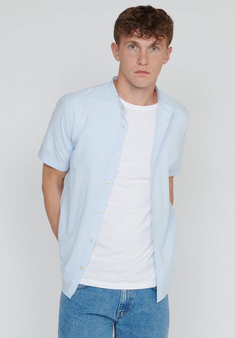 Jonge man met kort krullend haar, gekleed in een lichtblauw gestreept kortom shirt over een wit T-shirt en blauwe jeans, staande voor een effen achtergrond.